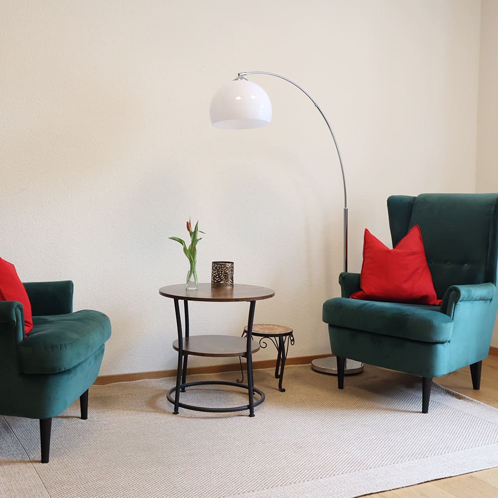 Zwei petrolfarbene Samt-Sessel mit je einem roten Kissen stehen auf einem hellen Teppich mit einem Tisch in der Mitte & einer Stehlampe vor einer hellen Wand