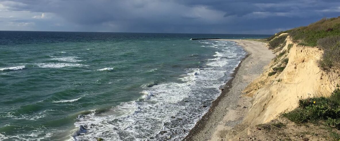 Küstenabschnitt mit Meer links, einem Steinstrand sowie Klippen rechts bei dunklen Wolken im Hintergrund