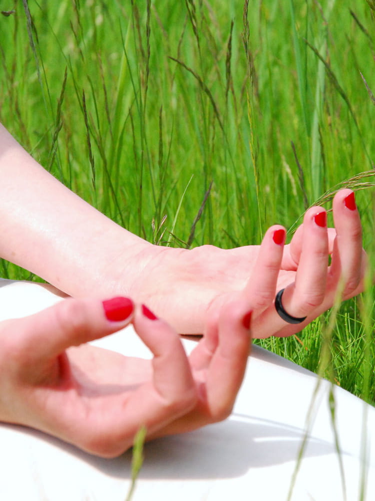 Zwei Hände mit rot lackierten Fingernägeln in einer Meditationspose auf einem weißen Kleidungsstück im grünen Gras sitzend