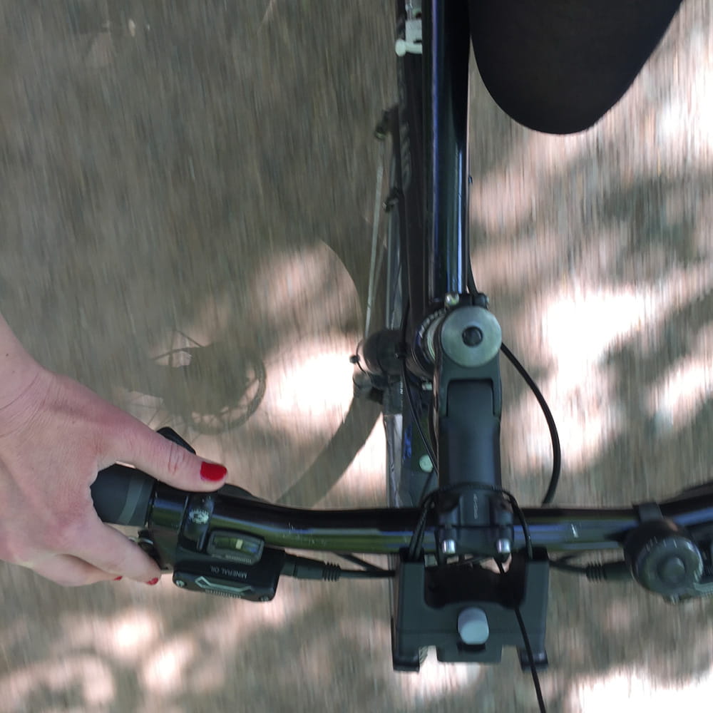 Blick nach unten auf eine auf einem Fahrrad sitzende Person mit einer Hand mit rot lackierten Fingernägeln am Lenker bei Sonnenschein