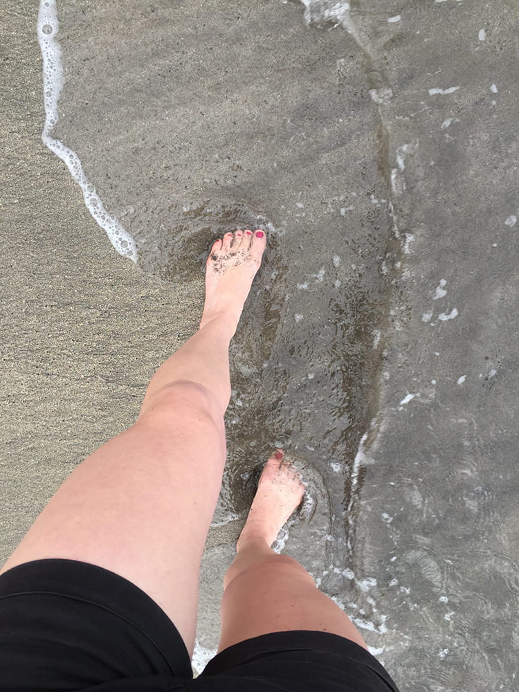 Blick nach unten auf Beine mit kurzer Hose & rot lackierten Fußnägeln & Meerwasser, das am Strand bis an die Füße läuft