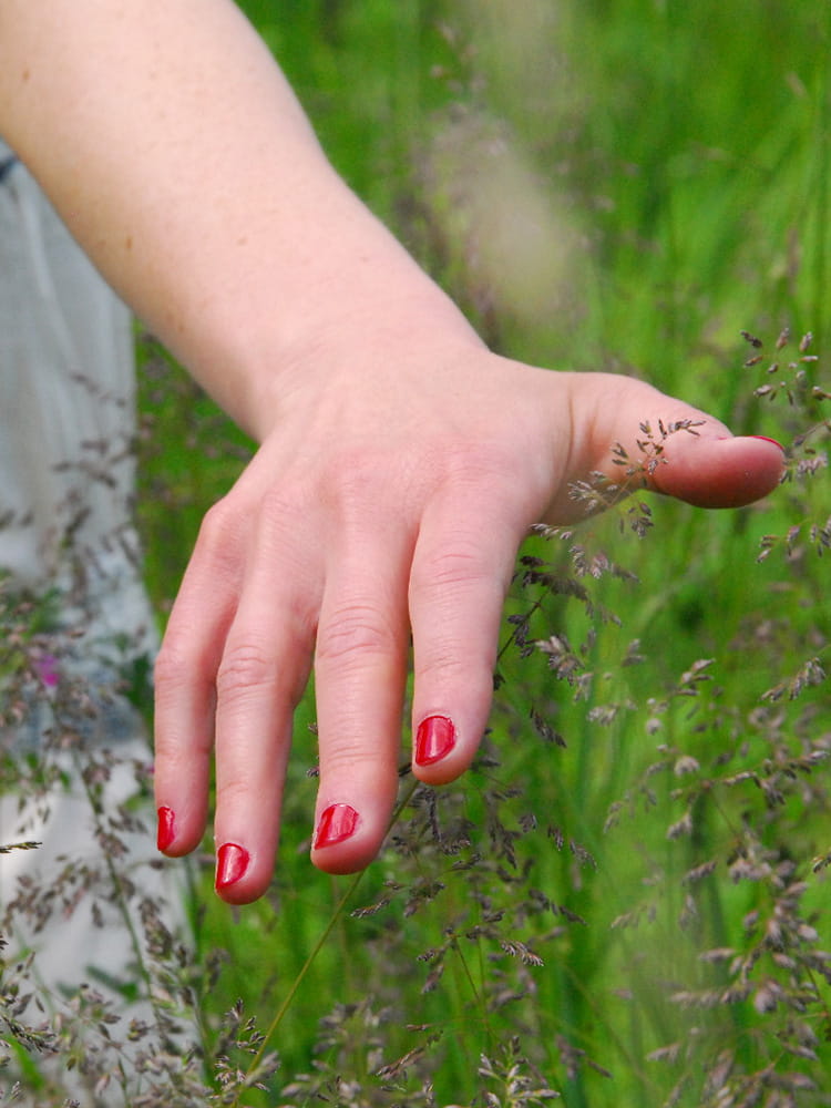 Hand mit rot lackierten Fingernägeln einer Person, die ein weißes Kleid trägt, fährt über Gräser