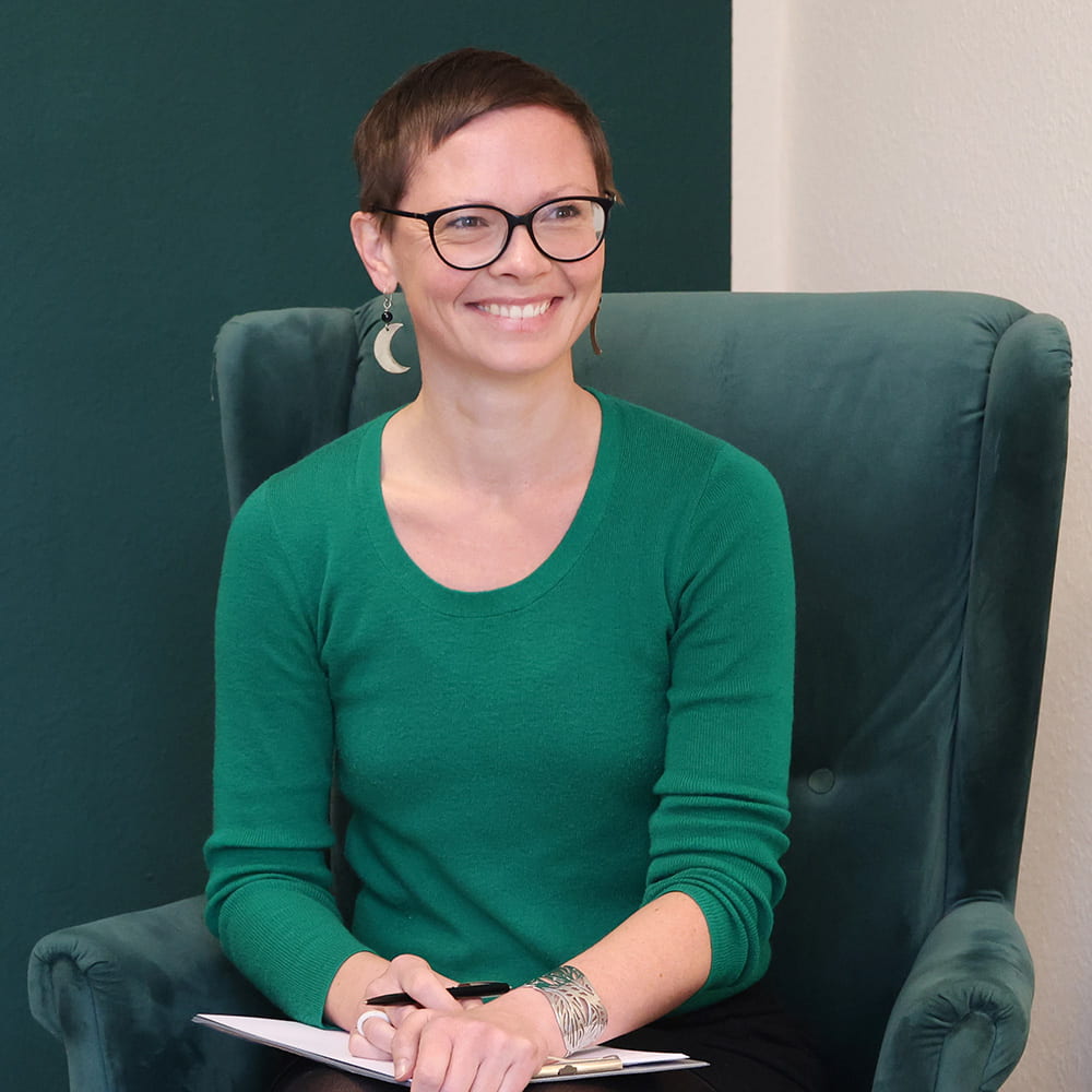 Dr. Carolin Peterhänsel sitzt in einem grünen Pullover mit Rundhalsausschnitt, einem Klemmbrett & einem Stift in der Hand, lächelnd auf einem petrolfarbenen Samt-Sessel vor einer petrolfarbenen & einer weißen Wand